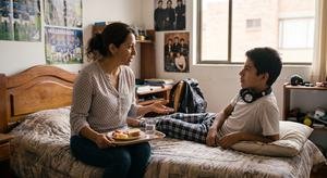 Una madre sentada al borde de la cama de su hijo adolescente, con una bandeja de comida en el regazo, le habla mientras él la mira.